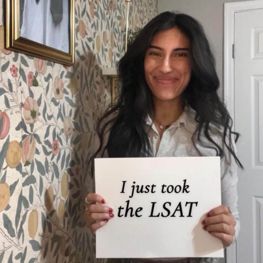 Una mujer se encuentra en el interior contra un papel pintado de flores sosteniendo un cartel en el que se lee "I just took the LSAT" (Acabo de hacer el examen de LSAT) con una sonrisa orgullosa en la cara.

