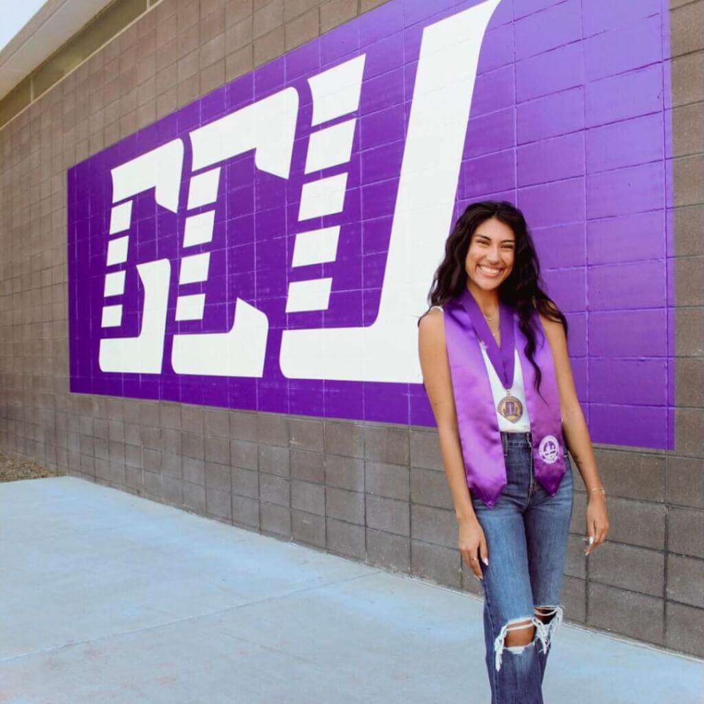 Una joven vestida con sus galas de graduación sonríe confiada delante de un mural morado que reza "GCU". Lleva una medalla y una estola de color lavanda.

