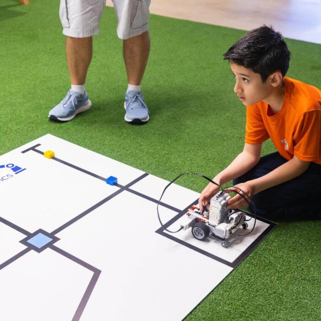 Un joven con camiseta naranja se arrodilla sobre césped artificial mientras maneja un robot con ruedas sobre una alfombrilla impresa. Cerca se ven las piernas de un adulto, lo que sugiere supervisión o instrucciones.