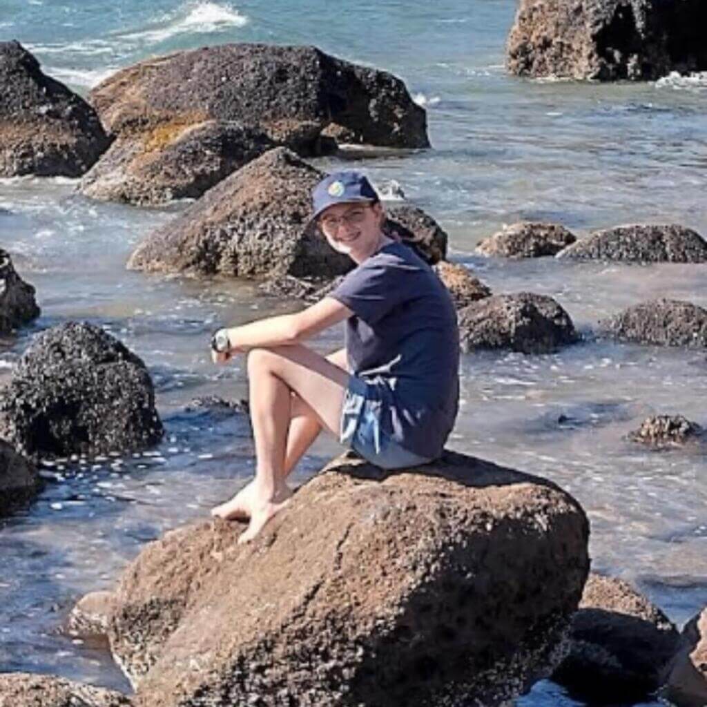 Un niño descalzo con traje azul marino y gorra está sentado en una gran roca rodeada de agua del océano y otras rocas, sonriendo mientras disfruta de un día soleado en la orilla.