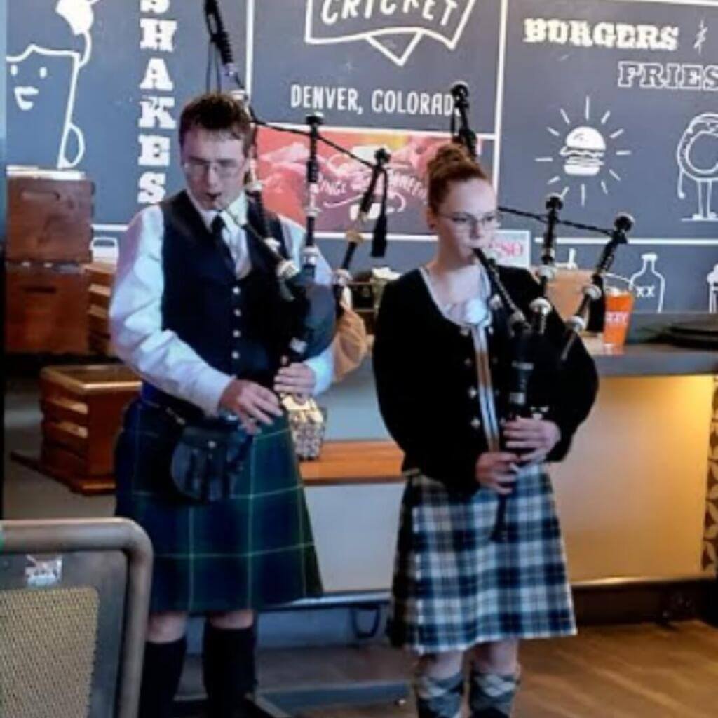Dos chicas y un chico vestidos con atuendos tradicionales de las Highlands sonríen para una foto en el interior. Las chicas llevan kilts de colores y chaquetas negras, mientras que el chico lleva chaqueta negra, kilt azul y boina.

