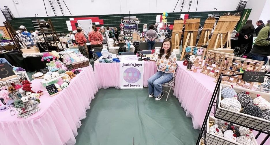 Puesto en una feria de artesanía con manteles rosas en los que se exhiben artículos hechos a mano, con el letrero «Janie's Joys and Jewels» (Las alegrías y joyas de Janie) y una persona sentada en el centro.