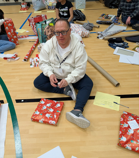 Una persona está sentada en el suelo de un gimnasio, rodeada de rollos de papel de regalo, cinta adhesiva y regalos envueltos, y está envolviendo un regalo con papel rojo con motivos navideños.