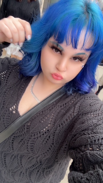 Una persona con el pelo de un azul brillante se hace un selfi vestida con un jersey negro y un collar.