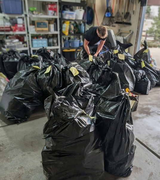 Una persona se inclina sobre un gran montón de bolsas de basura negras llenas en un garaje, pegando o revisando las etiquetas de las bolsas. En la pared del fondo hay estanterías con objetos almacenados.