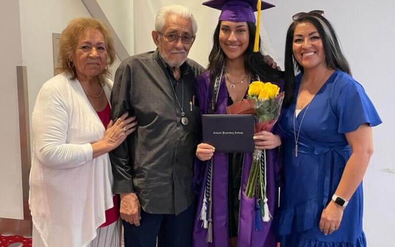 Un graduado con toga y birrete morados sostiene un diploma y flores amarillas junto a tres familiares sonrientes, entre ellos dos mujeres y un anciano.