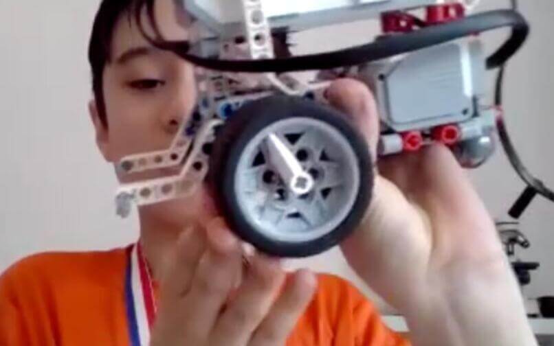 Un niño con una camiseta naranja y una medalla sostiene un robot estilo LEGO, mostrando de cerca su rueda y su cableado, exhibiendo orgulloso su proyecto.