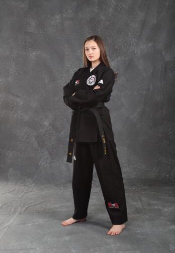 Persona descalza con un uniforme negro de artes marciales y cinturón negro, con los brazos cruzados, sobre un fondo gris de estudio.