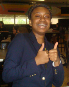Persona con una chaqueta azul marino haciendo un gesto de aprobación con los pulgares en una cafetería interior.
