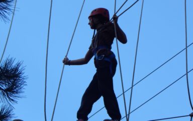 Un participante, equipado con casco y arnés de seguridad, cruza un puente colgante de cuerdas en un circuito de cuerdas al aire libre.