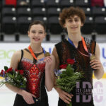 Dos patinadoras artísticas, con medallas de oro colgadas al cuello, sostienen ramos de flores y posan sobre el hielo tras una competición.