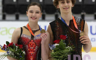Dos patinadoras artísticas, con medallas de oro colgadas al cuello, sostienen ramos de flores y posan sobre el hielo tras una competición.